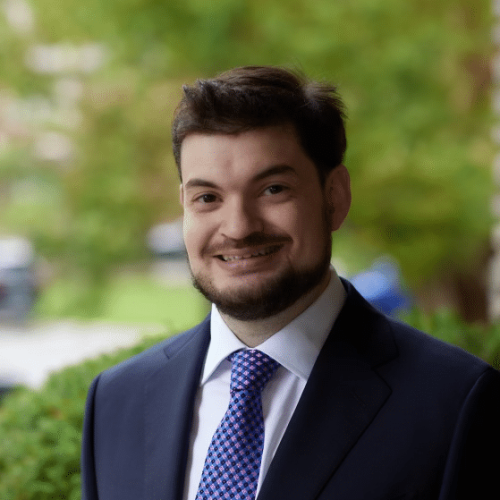 A man with short dark hair, a trimmed beard, and mustache, wearing a dark suit, white shirt, and patterned tie, smiles outdoors with greenery behind him—portraying the confidence of a Copyright Lawyer Toronto professional.